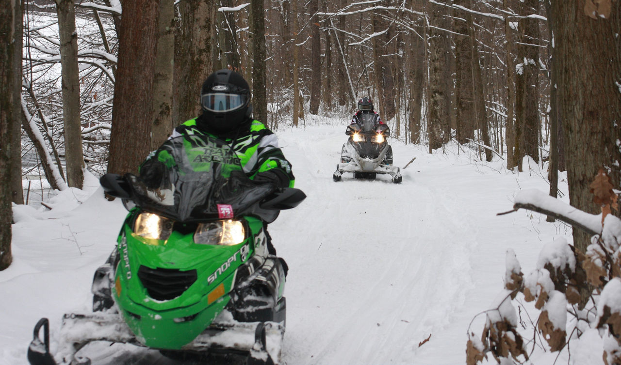 Two snowmobiles driving on a wide snowy path through the forest