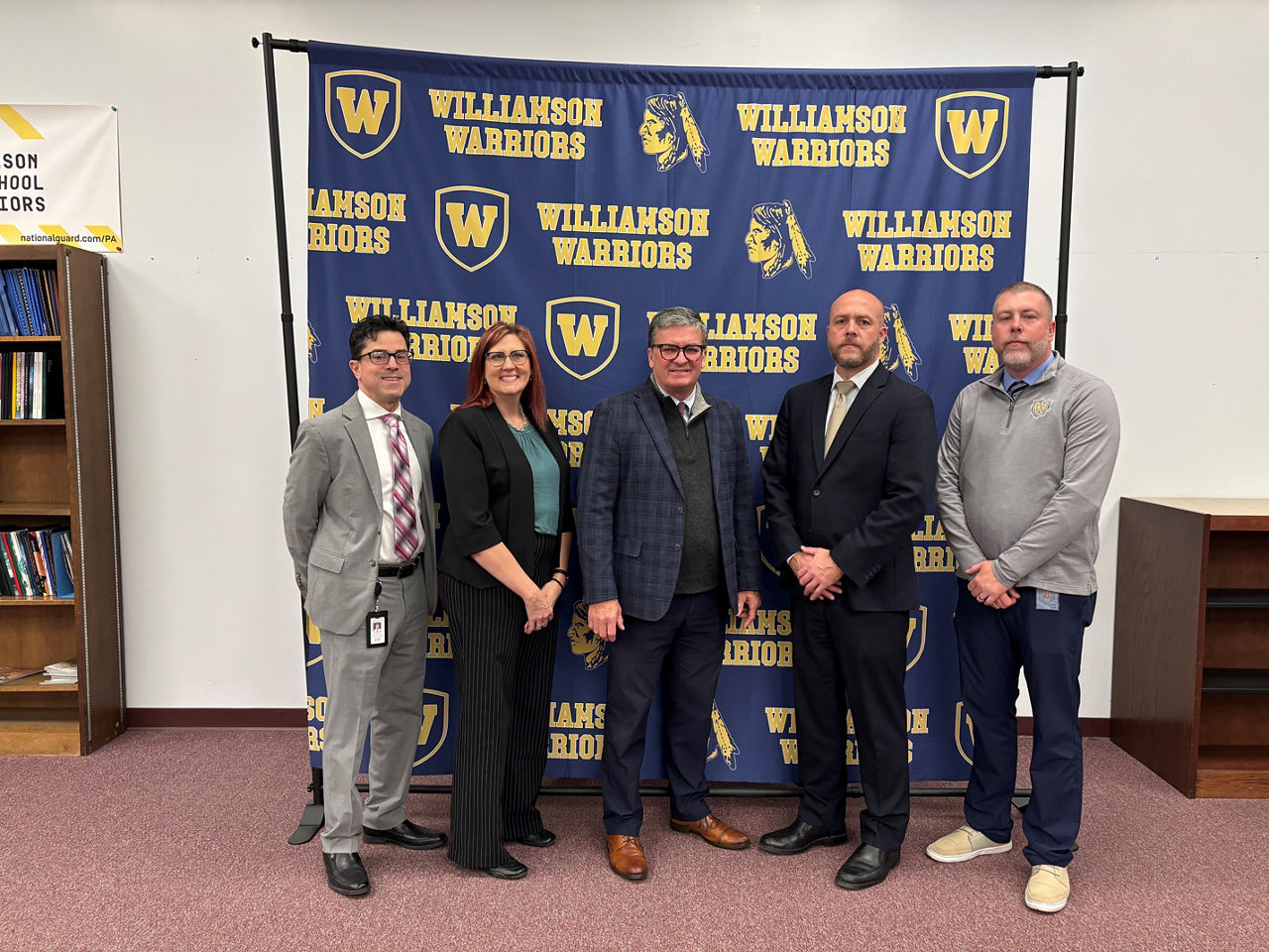  Four men and one woman stand in front of a photo backdrop displaying the Williamson High School name and mascot.
