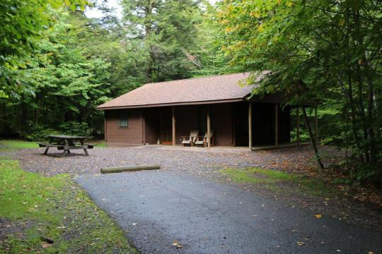 A small wooden cabin nestled in the woods with a ramp leading up to the front porch, with a fire ring and picnic table visible in the yard