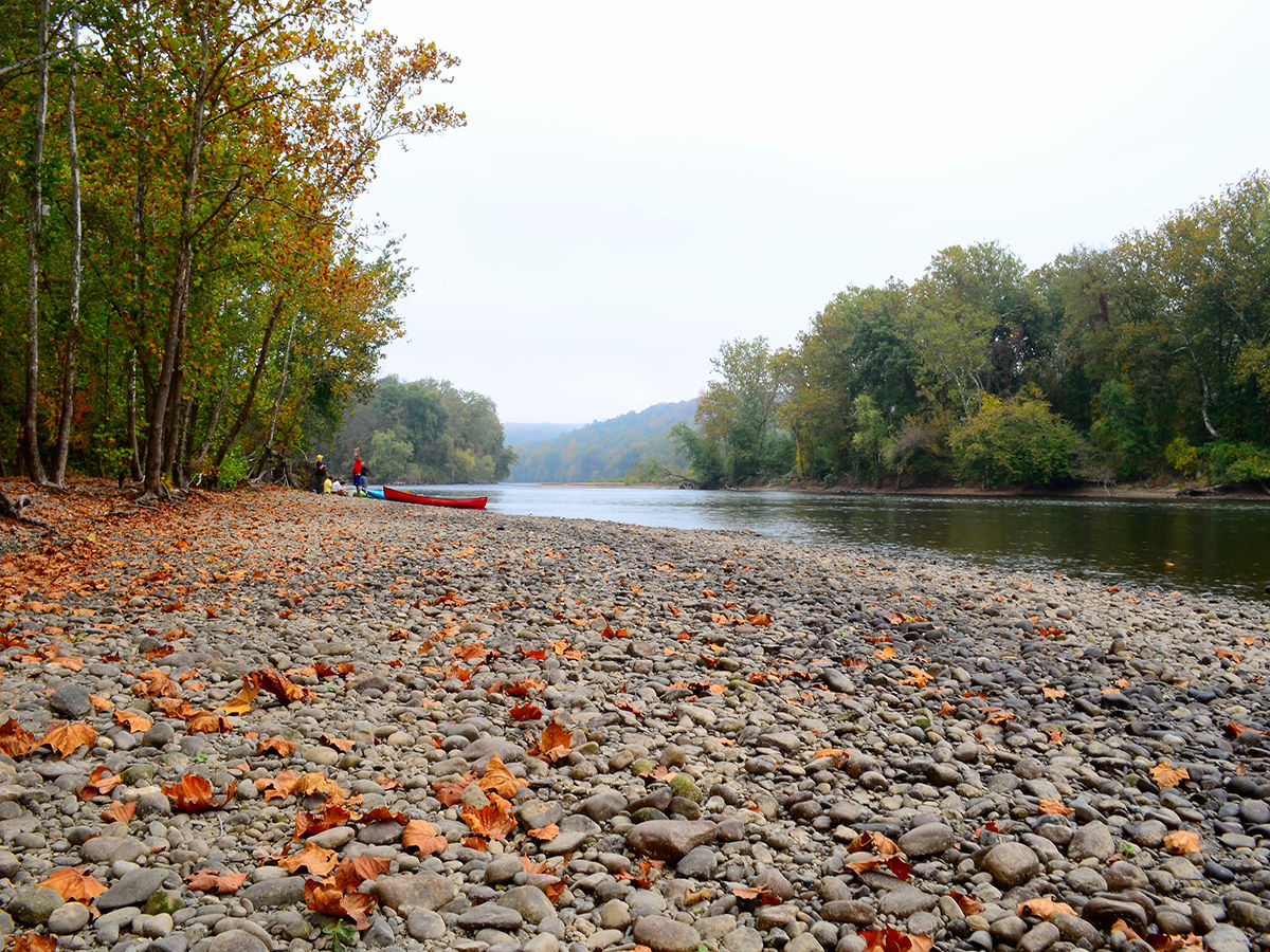 Rocks line the ground next to the Delaware River. A group of people appear by a boat in the background.