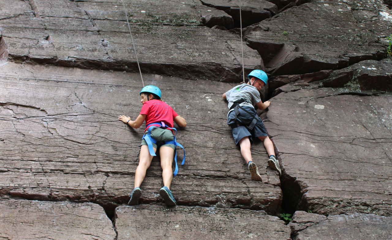 Two youth climbing up a cliff face wearing climbing equipment