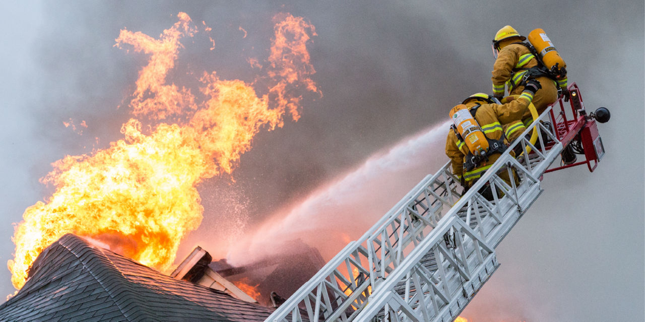 Firefighters on a ladder blast fire coming out of a roof with water.