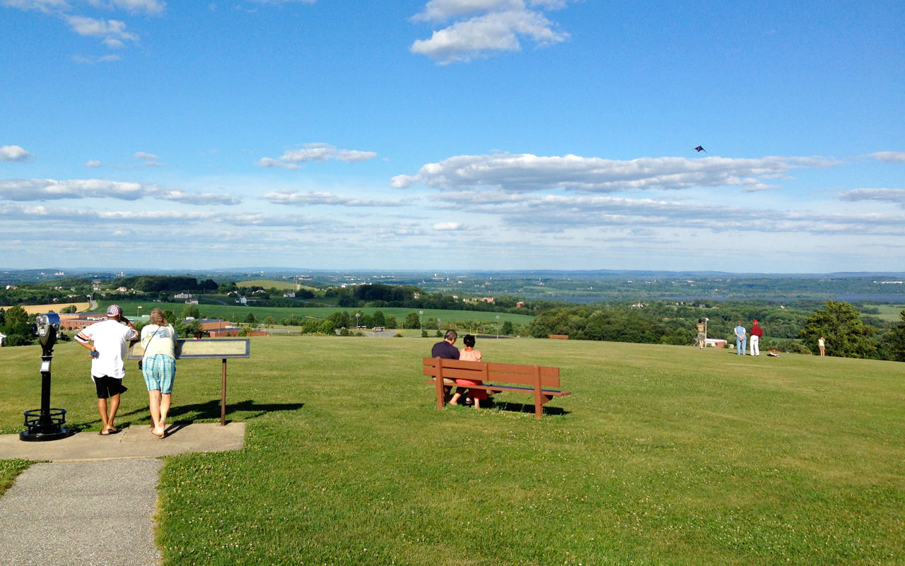 A couple at an overlook at the end of a path. Nearby a couple sits on a wooden bench in a grassy area.