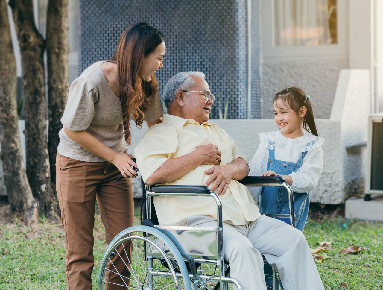 Happy family - man in wheelchair being pushed outdoors with daughter and granddaughter