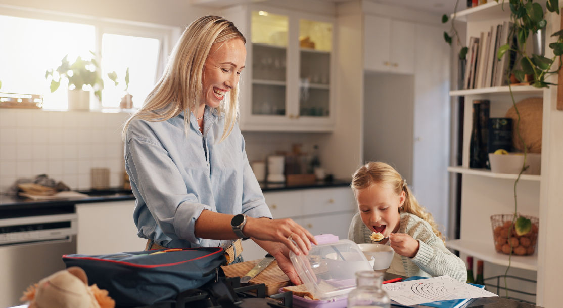 A mom preps her daughters lunch while she eats breakfast before school.