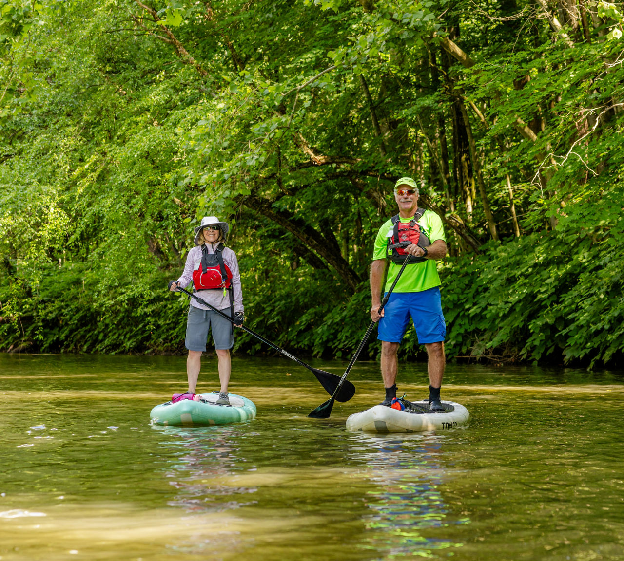 A man and a woman, both wearing life jackets, paddleboard along the Schuylkill River.