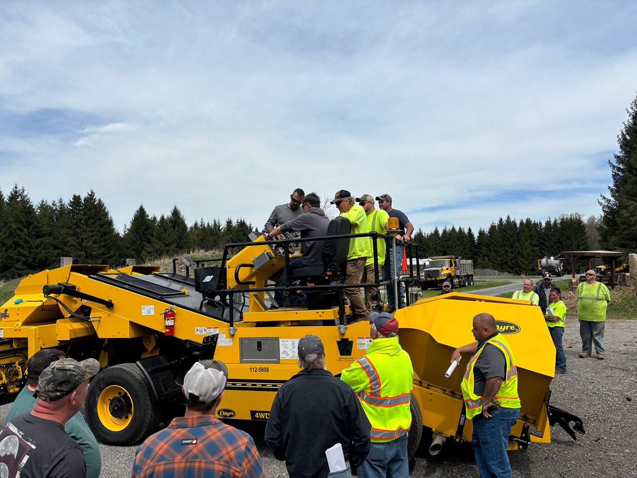 Several roadway maintenance crew members stand around and atop a large of piece of equipment as an instructor demonstrates how to operate the piece of equipment.