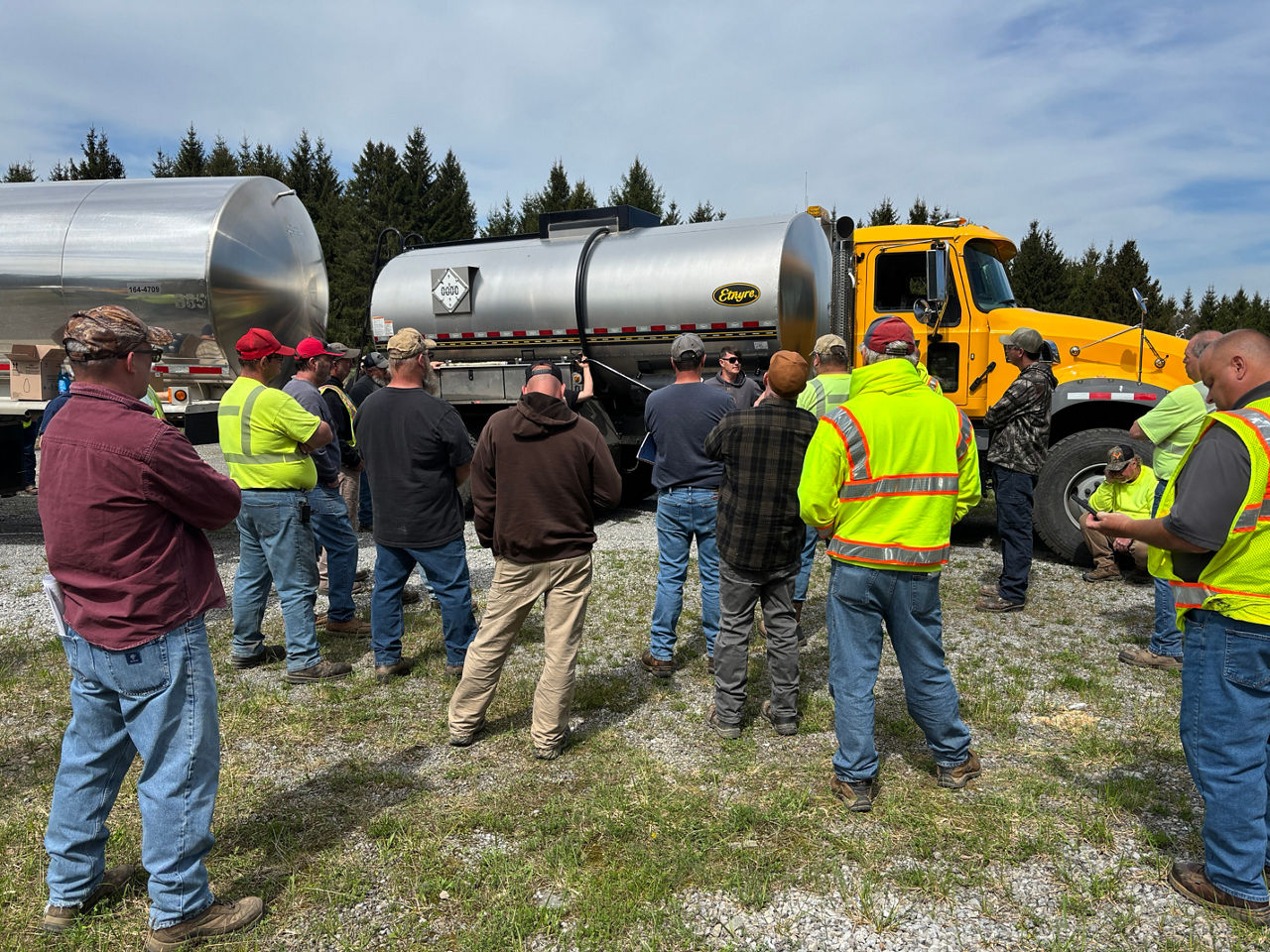 Several roadway maintenance crew members stand in front of an oil distributor truck listening to an instructor discuss proper operation of the equipment. 