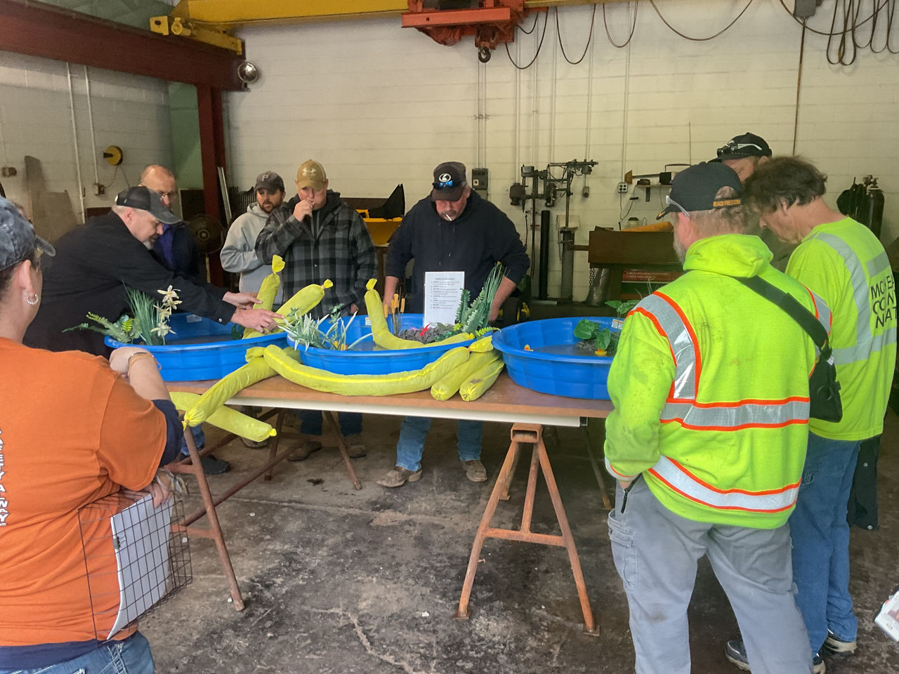 An image of PennDOT employees standing around three blue baby pools filled with water that sit atop a table. Each pool is filled with rock, artificial plants, toy animals and insects, and a prop oil-drum. 
