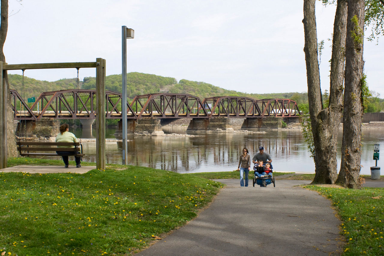 A couple pushing a stroller along a paved path beside the river. A woman on a bench swing overlooks  the river and metal bridges