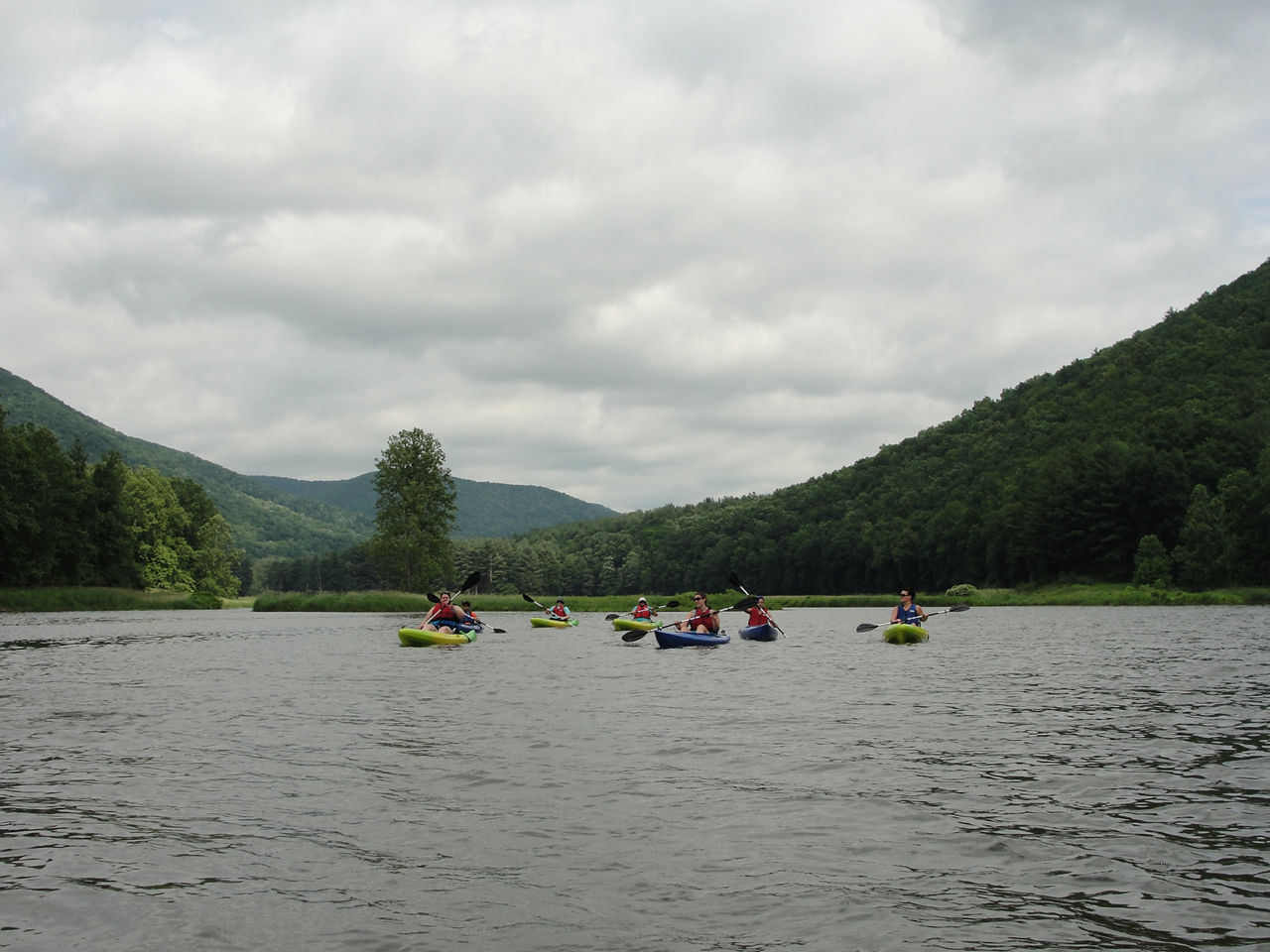 On a cloudy day, a group of kayakers paddle in the lake surrounded by forested mountains