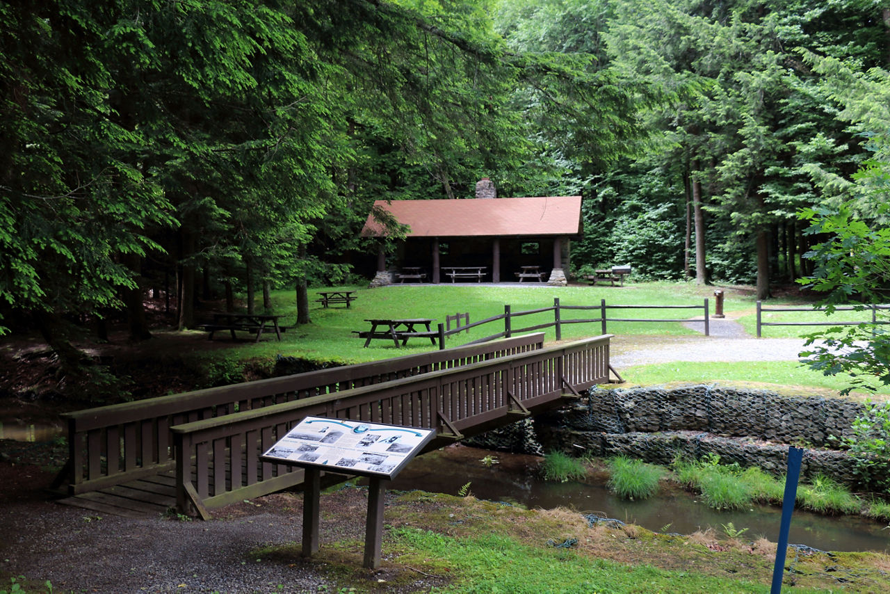 An information sign is beside a wooden bridge crossing a stream to the day use are on the other side