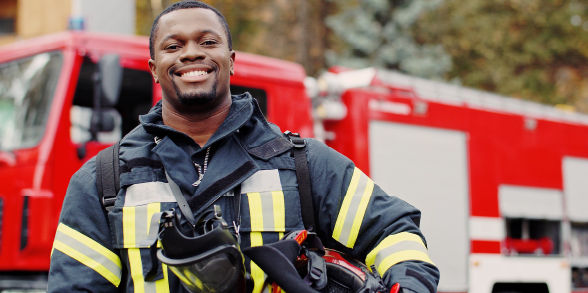 A Black male firefighter smiles at the camera while holding his helmet in front of a fire truck.