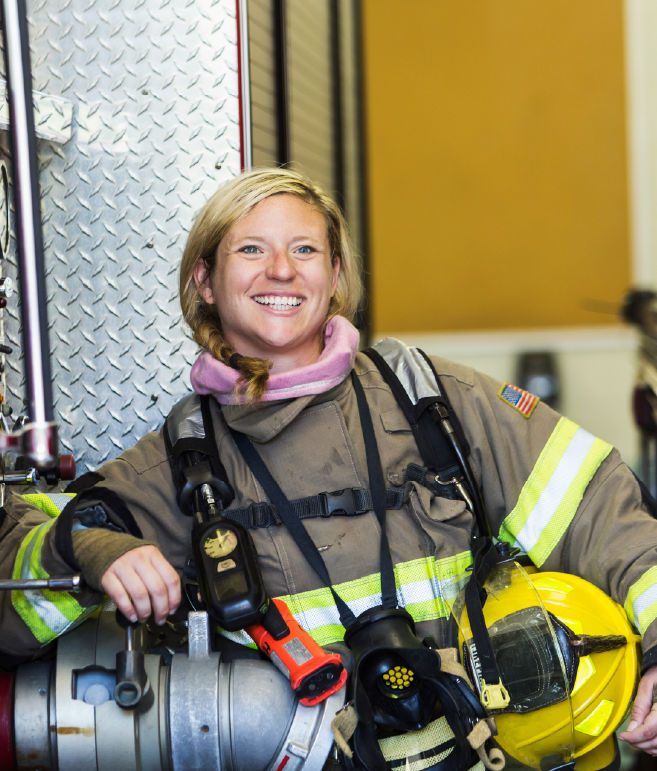 A blond, female firefighter smiles while leaning up against a firetruck.