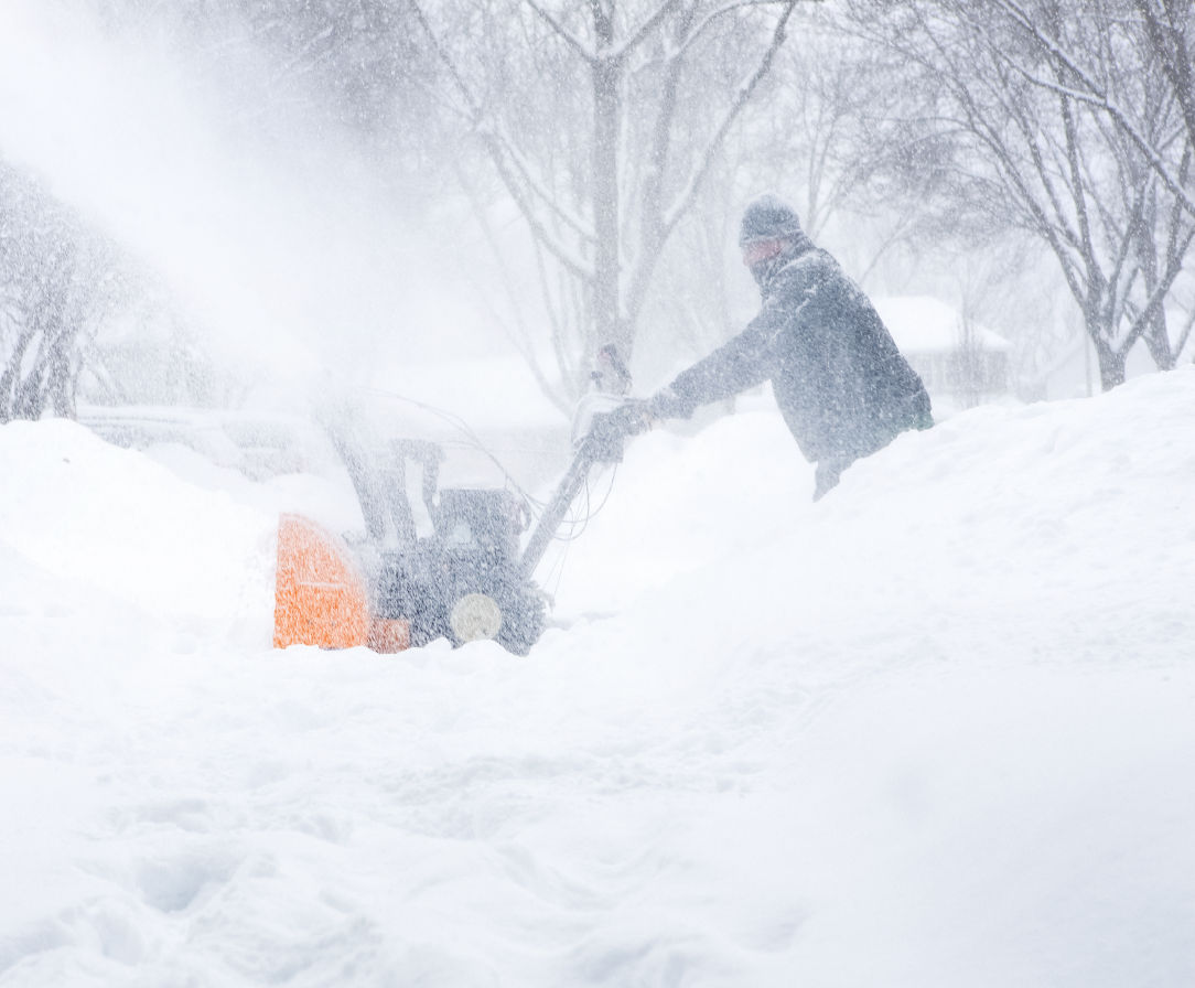 A person bundled up in winter attire snowblows several feet of snow.