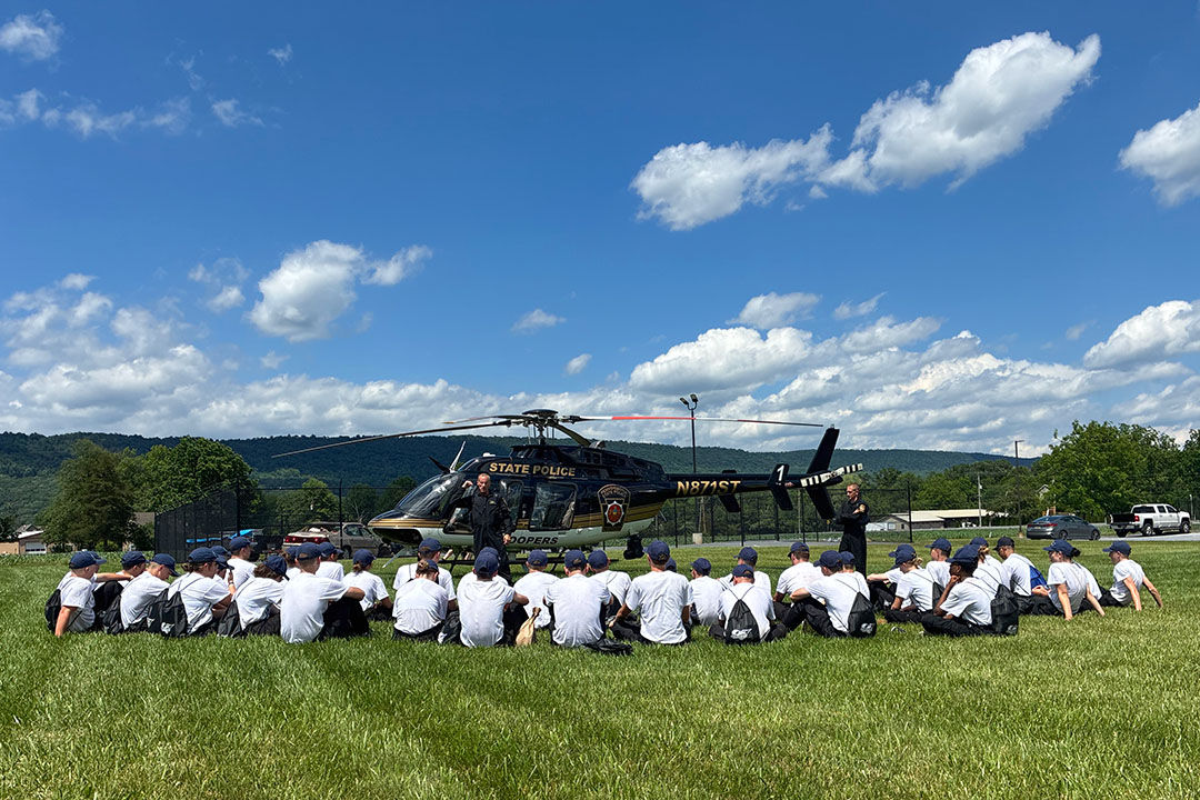 On a sunny day, a group of youths sit near a State Police helicopter in a grassy field while two pilots address the group