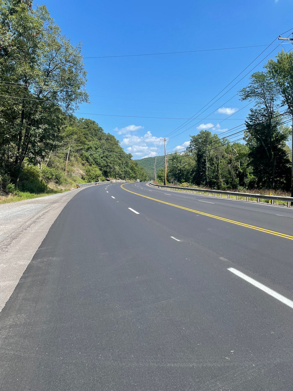 A white truck approaches on a tree-lined, four-lane roadway divided by a double yellow line.
