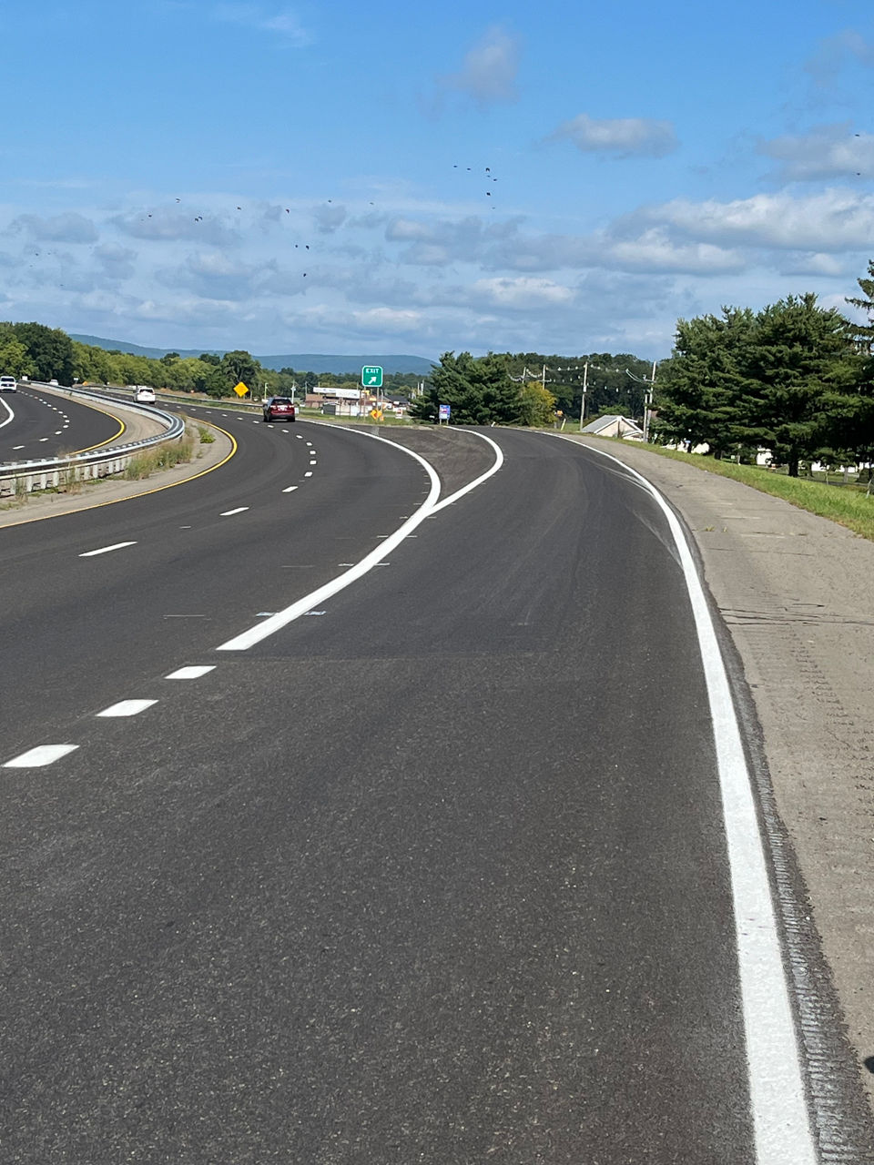 A red car is headed northbound on a tree-lined, four-lane roadway divided by a guardrail and grassy area.