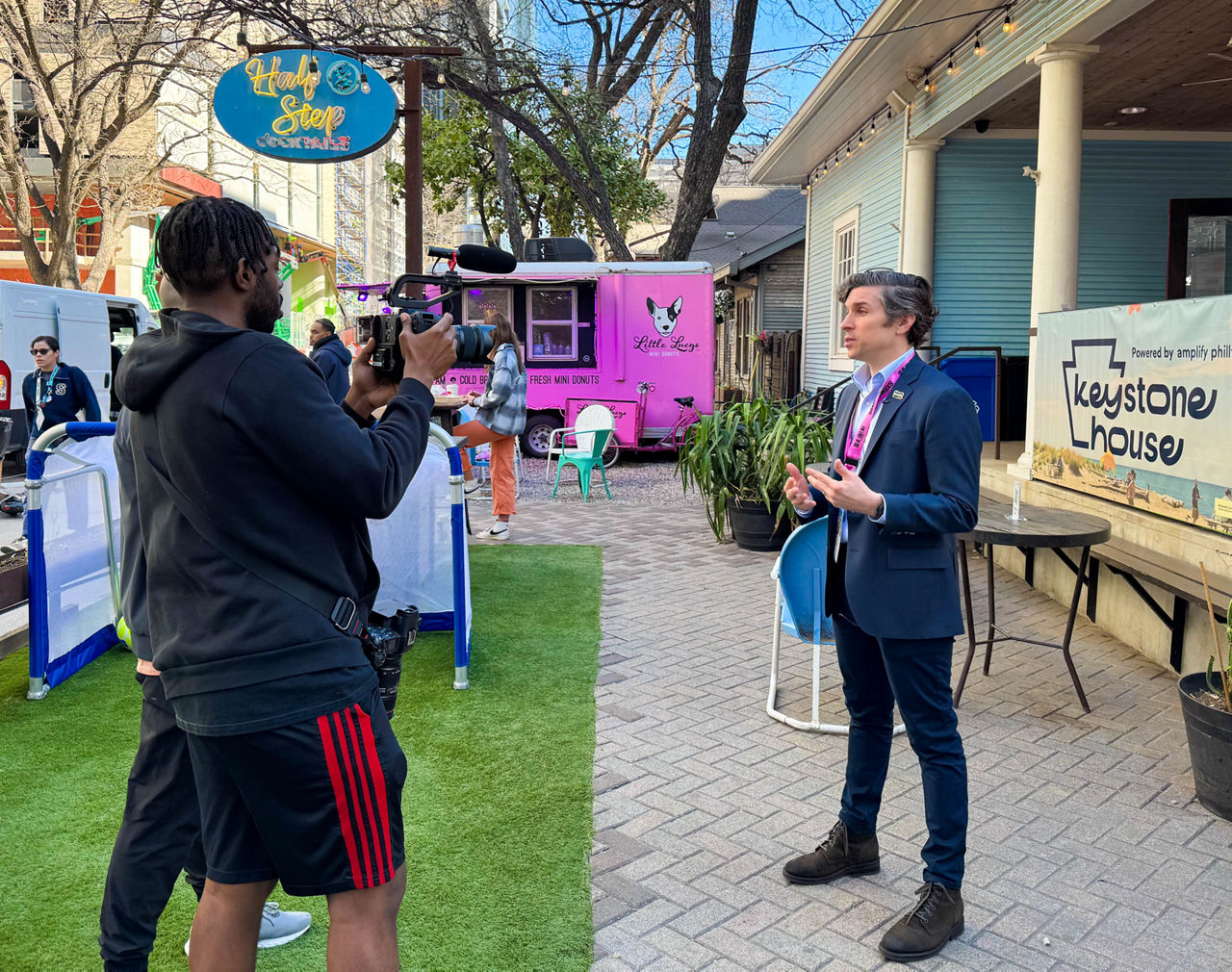 Michael O'Rourke is interviewed by two videographers at an outdoor event at SXSW. A food truck and crowd are visible in the distance.