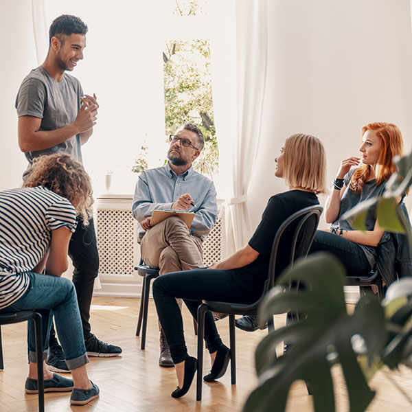 A counselor leading a group therapy session with a group of teens. One teen is standing and smiling while sharing with the group.