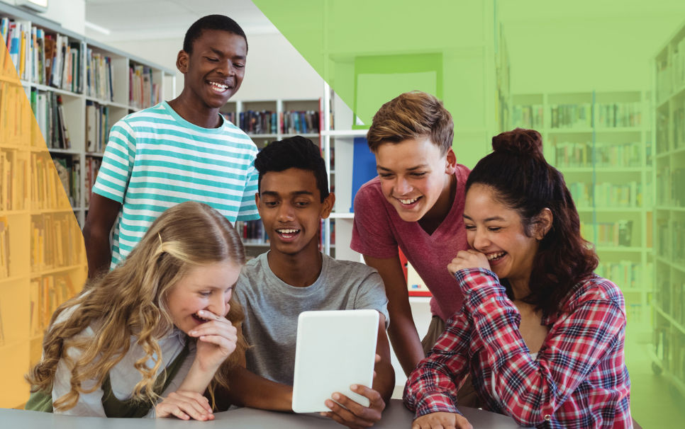 a group of teenagers laughing around a tablet in a library