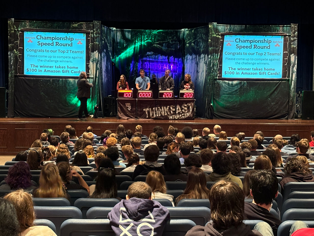 A large high school auditorium filled with adults and students. A stage is up front with a table and chair in the center and two large screens on either side of the table. Four students stand behind four monitors displaying their game show scores.