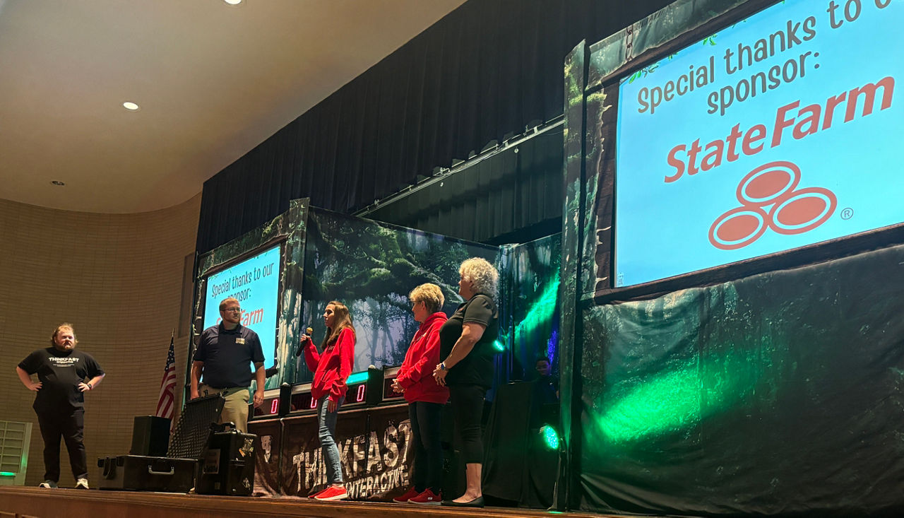 A student is speaking into a microphone on a large auditorium stage with two adults standing on either side. Behind these individuals are a table and two large screens on either side of the table.