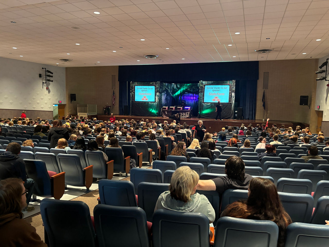 A large high school auditorium filled with adults and students. A stage is up front with a table and chair in the center and two large screens on either side of the table.