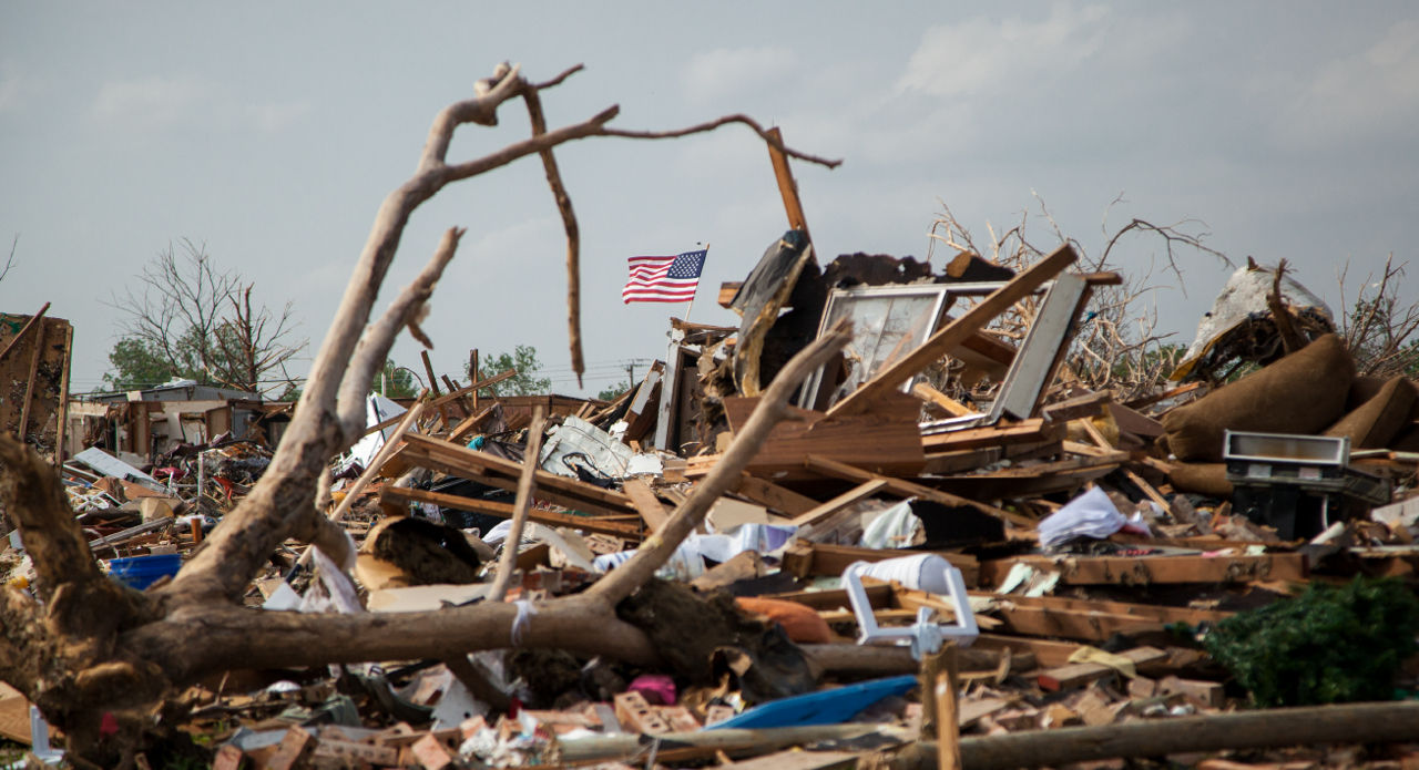 An American flag stands tall over a house that has been reduced to rubble.