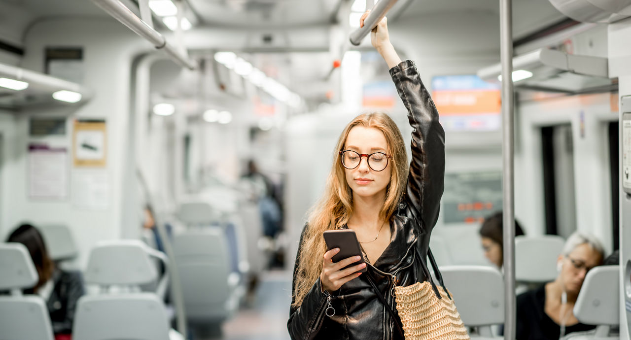 A young woman wearing glasses and a leather jacket checks her phone while riding a train.