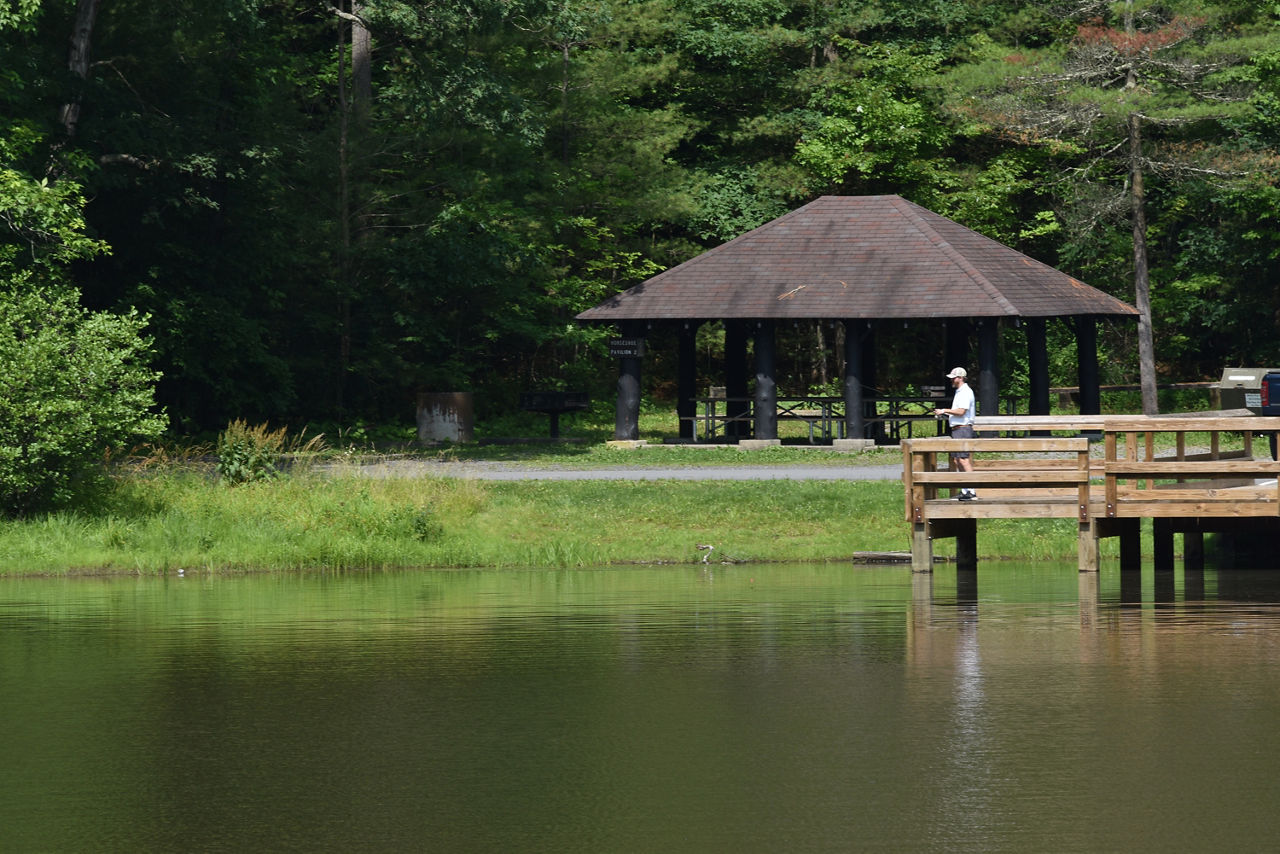 A fisherman at the end of a pier over a small lake with a pavilion in the background