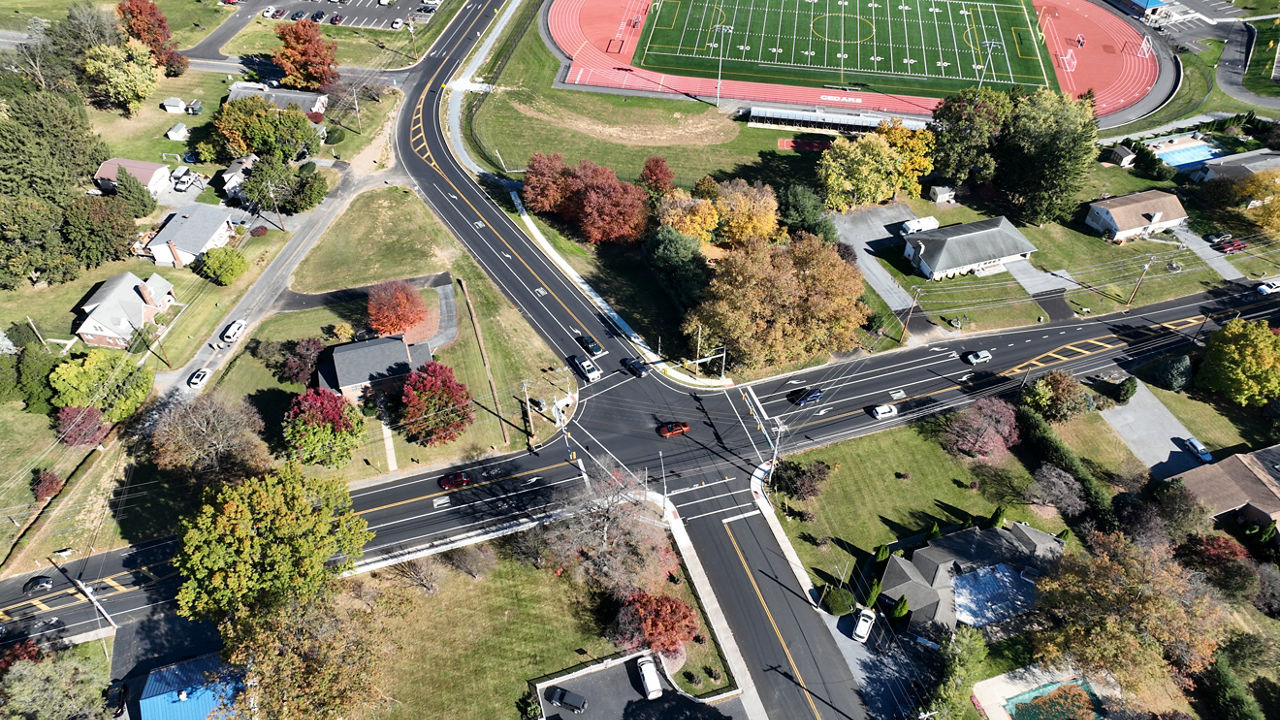 An aerial view of two intersecting roadways surrounded by houses and trees and open grassy areas. A football field sits in the background of the photo.