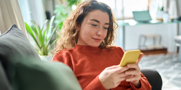 A girl with red, curly hair lounges on a couch in her living roo while staring down at her phone.