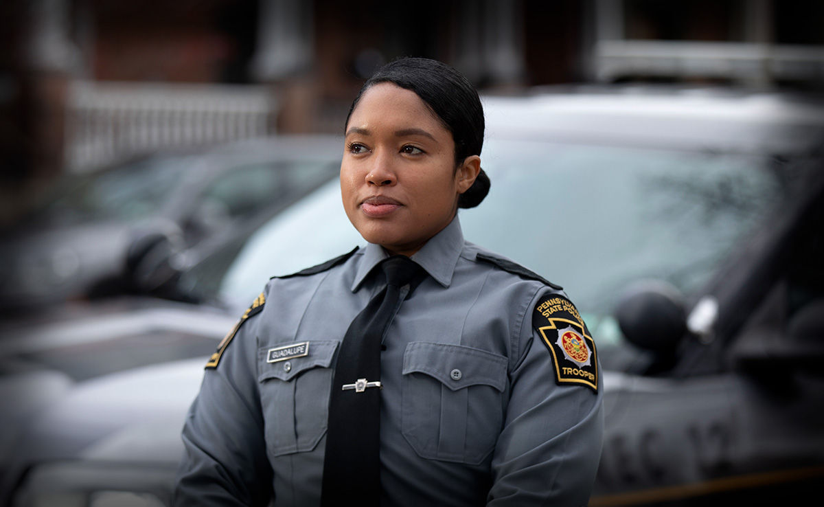 A female state police Trooper in front of a marked state police vehicle