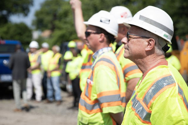 Workers stand in a row. They are wearing yellow safety vests and white hardhats, listening to someone offscreen speak