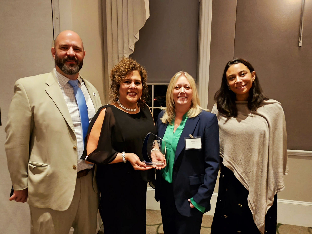 Three woman and one man standing next to each other in front of a wall and window. One of the woman holds a glass trophy in her hands.
