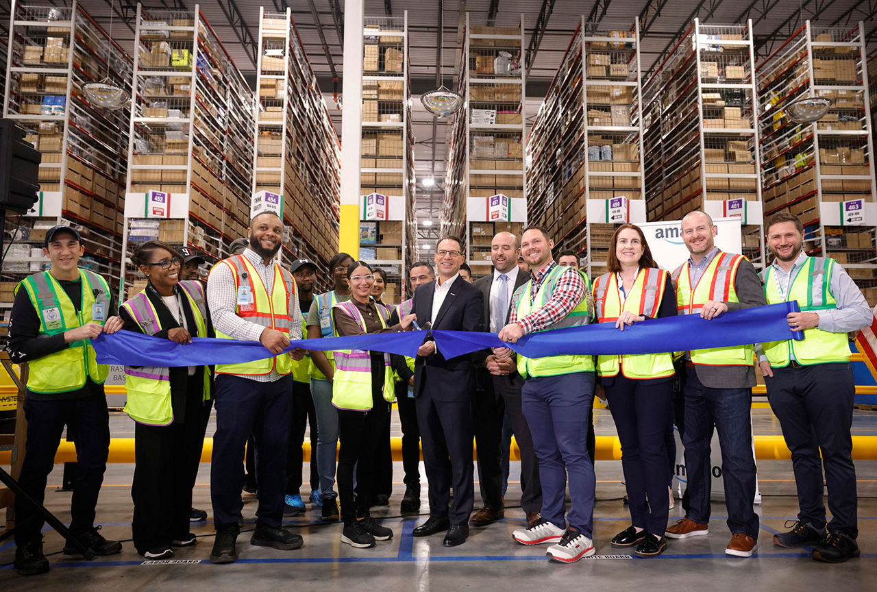 Governor Josh Shapiro cuts a blue ribbon with help from United Parcel Service (UPS) employees, in a warehouse filled with shelves stacked high with shipping boxes.
