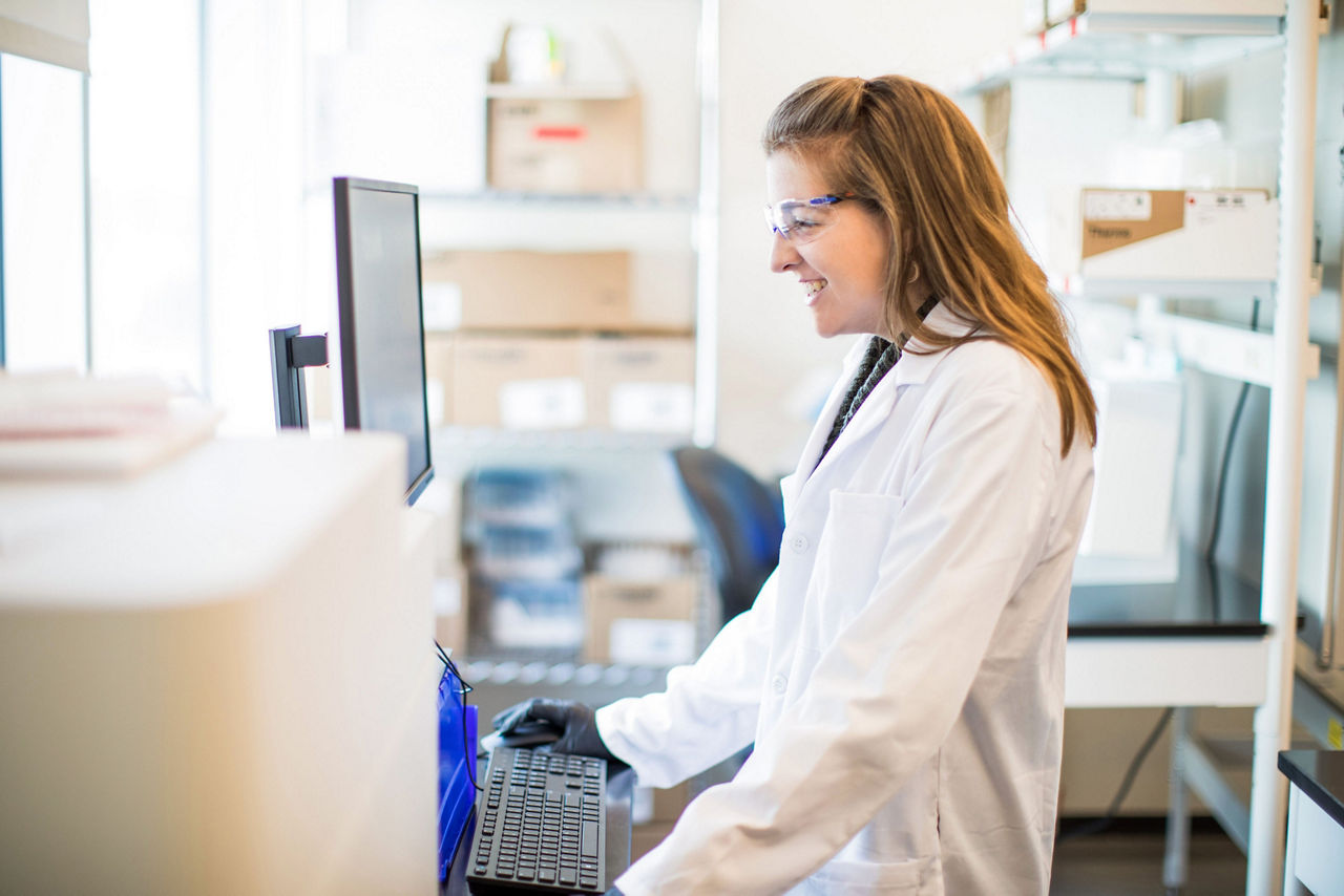 A smiling scientist wearing a lab coat works at her computer in a bright laboratory