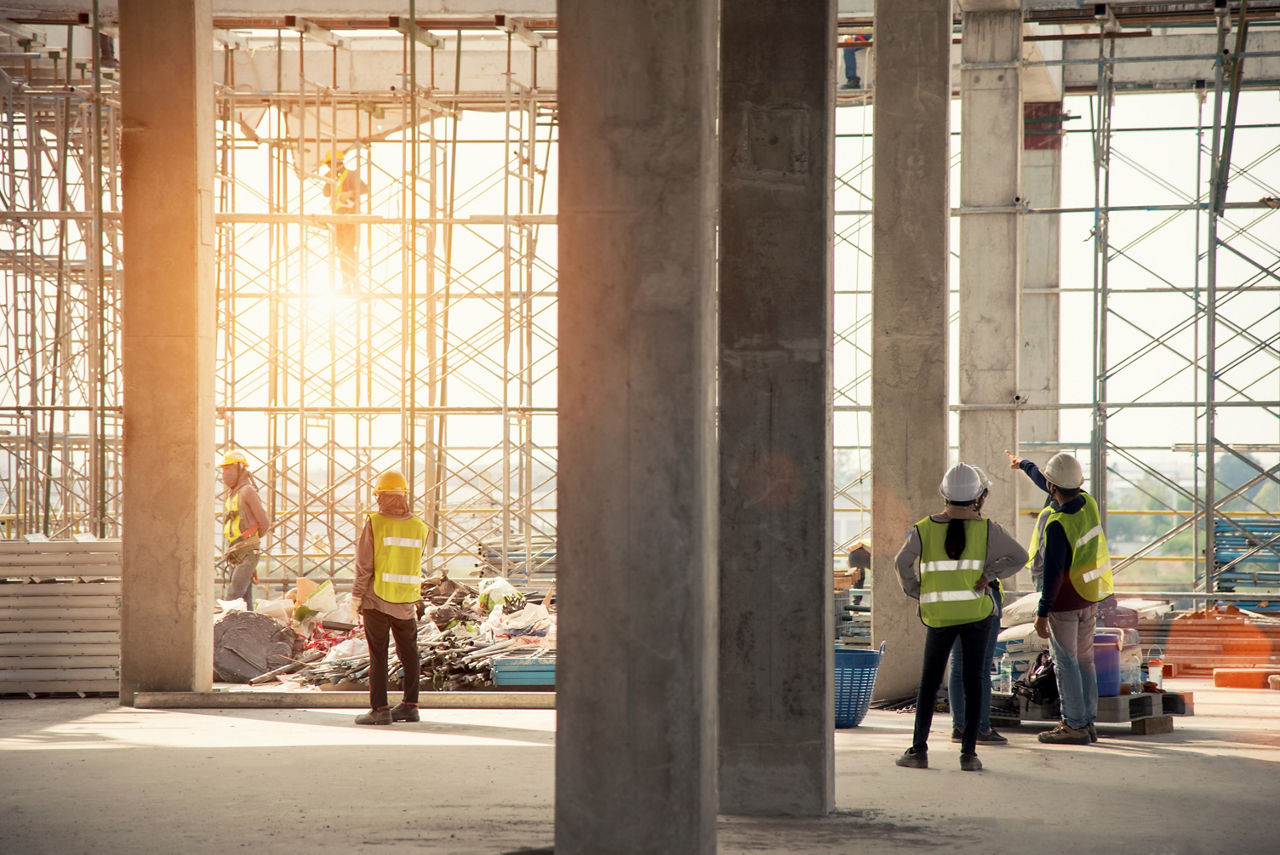 Construction workers inspect the inside of an under-construction building. A sunset is visible through scaffolding in the background.