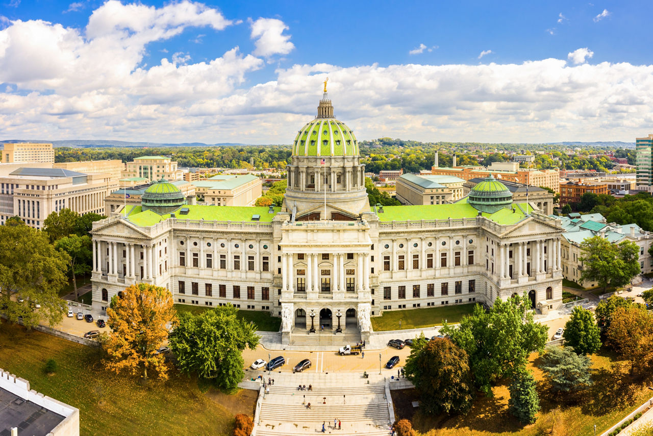Aerial view of the Pennsylvania state capitol building, an ornate stucture surrounded by trees, with Harrisburg visible in the background.