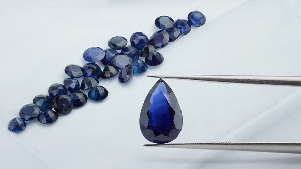 Tweezers holding a blue gemstones with more gemstones on the table 