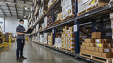 Male wearing a mask and using a laptop in a warehouse full of boxes