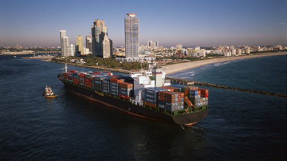 Cargo ship full of shipping containers going through water next to a city on the coast