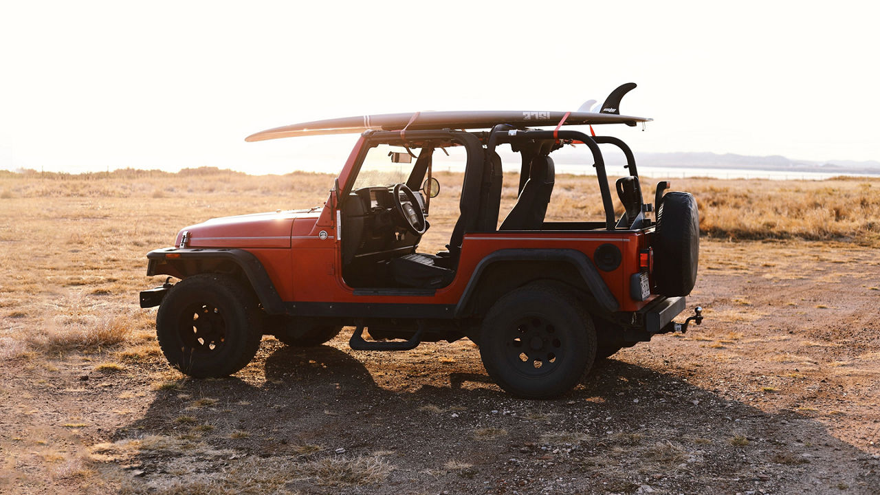 Red Jeep on the dirt with no doors and a surfboard