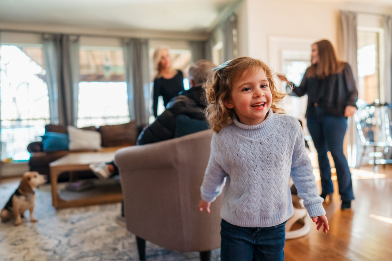 A young child stands in the foreground of a warmly lit living room, wearing a light sweater and jeans. In the background, adults are engaged in conversation, and a small dog sits near a coffee table. The scene suggests a casual family gathering in a comfortable home environment, with natural light streaming through large windows.
