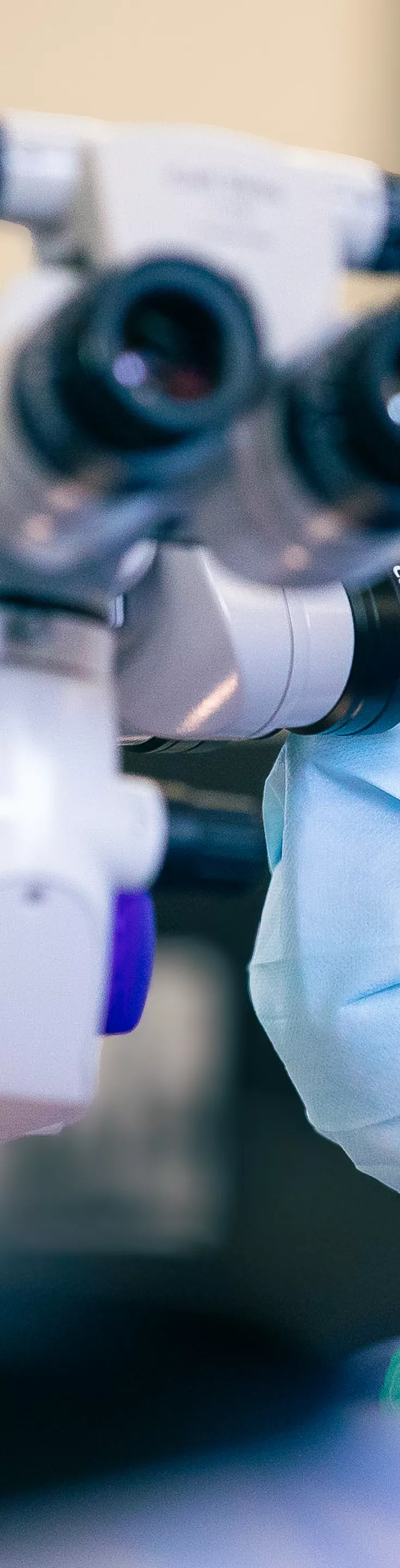 A medical professional is closely observing through a surgical microscope in a clinical setting. The individual is wearing a red surgical cap and a face mask, indicating a sterile environment. The background is softly blurred, focusing attention on the advanced optical equipment and the surgeon's concentration. 