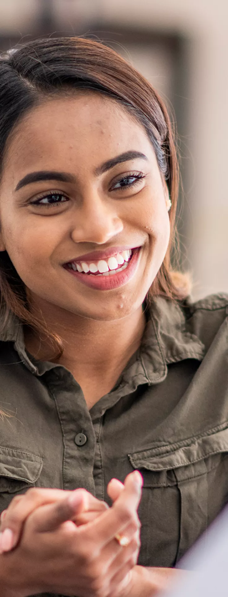 A young woman smiles at another woman with her back to the camera. She is wearing a green button up shirt and has her hands clasped together. They're seated in a softly lit room.