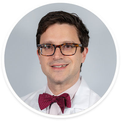 A male adult is pictured wearing a white medical coat and a red polka dot bow tie. The background is a plain, light gray, and the subject is centered in a circular frame. The attire and setting suggest a professional or clinical environment, emphasizing a formal and polished appearance.