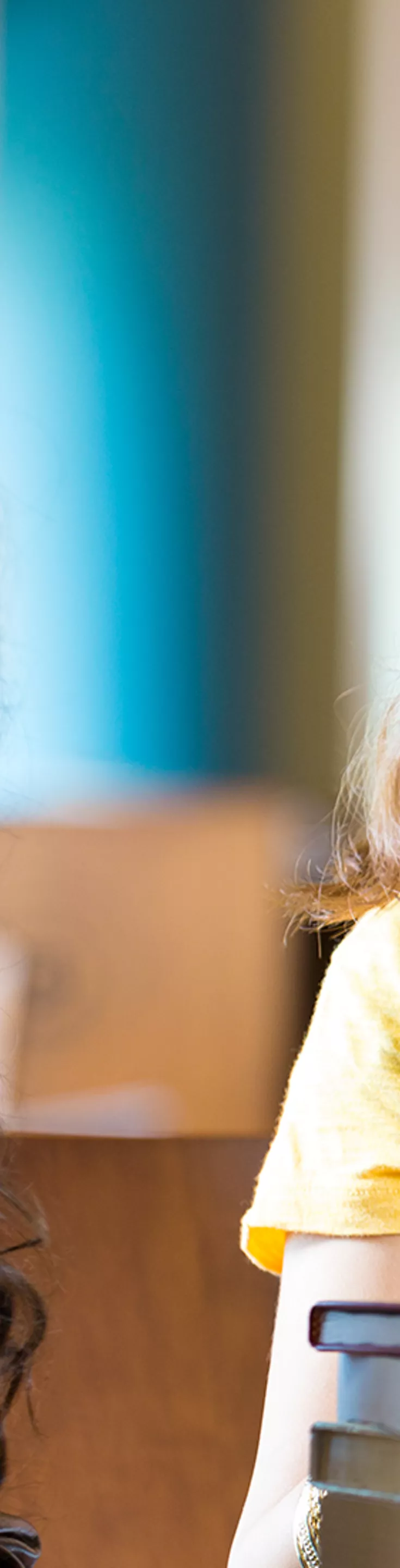 A young woman is smiling at another person who has their back to the camera. One hand is on her chest and the other rests on top of a stack of books. She is wearing a yellow shirt, black glasses, and a cream colored vest.