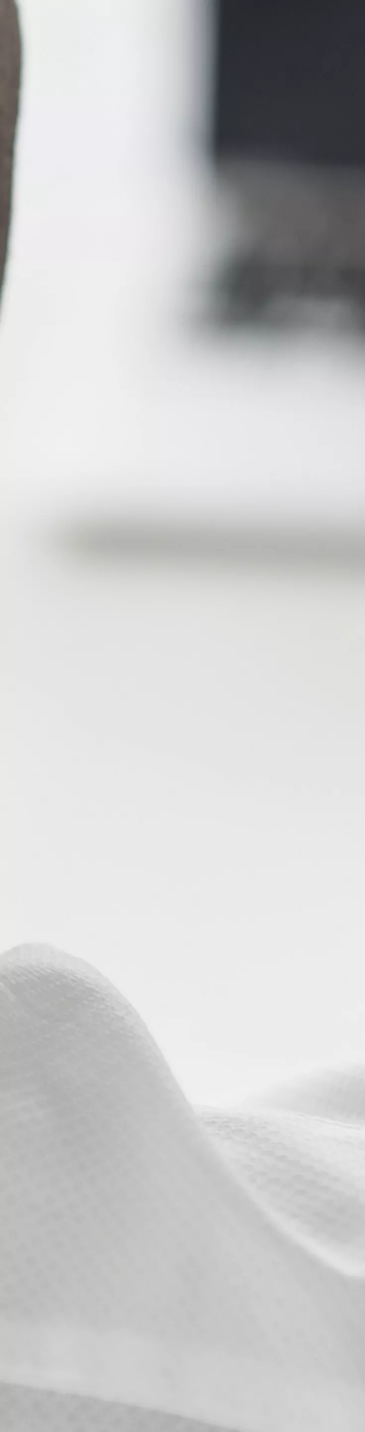 A man in a white shirt is seen from behind, holding his neck as if experiencing discomfort. He is seated at a desk with a smartphone in one hand and a laptop in the background. The setting suggests an office environment, with a focus on workplace stress or fatigue. The overall palette is neutral and professional.